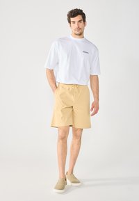 Beige shorts met een elastische tailleband en trekkoord, gecombineerd met een wit t-shirt met zwarte tekst. Lichtgekleurde instappers.