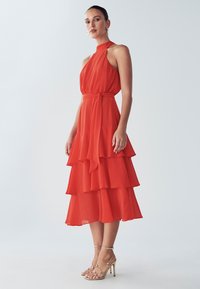 Rode chiffonjurk met halternek, gelaagde tiers en een taillekoord. Gecombineerd met gouden sandalen met hoge hak en bandjes. Zachte val en beweging.