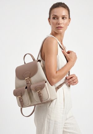 Vrouw draagt lichtbeige mouwloos kledingstuk en draagt een beige canvas en leren rugzak met drie voorvakken met gespen.