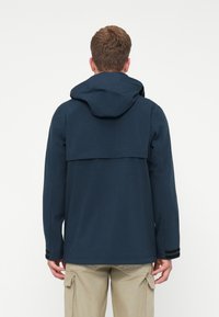 Άνδρας που φοράει μπλε φούτερ με κουκούλα και μπεζ cargo παντελόνι, στέκεται με την πλάτη στραμμένη προς τα εμπρός σε λευκό φόντο.