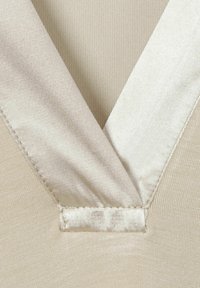 Beiges Kleidungsstück mit V-Ausschnitt und Satinbesatz. Die Textur ist glatt, und es sind sichtbare Nähte entlang der Kanten vorhanden.