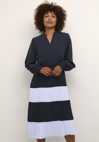 Tumši zila kleita ar garām piedurknēm, plisēta apakšējā daļa ar maiņus tumši ziliem un baltajiem svītrām, un V-veida dekoltē.