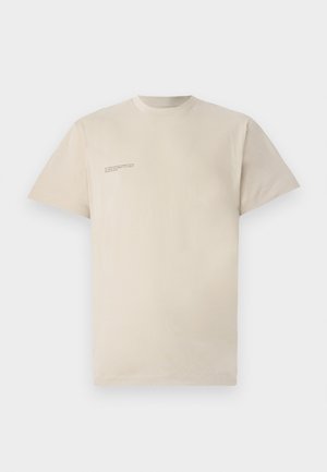 Camiseta de manga corta beige en tejido suave. Presenta un cuello redondo y una pequeña etiqueta impresa en el área del pecho izquierdo. Diseño minimalista.