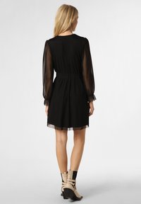 Schwarzes Kleid mit durchsichtigen Puffärmeln, elastischem Bund und geschichtetem Saum. Das Modell trägt beigefarbene Stiefeletten mit schwarzen Akzenten.