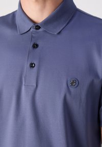 Polo tričko z fialové látky s klasickým límečkem, třemi černými knoflíky a kulatou nášivkou na levé straně. Hladká textura.