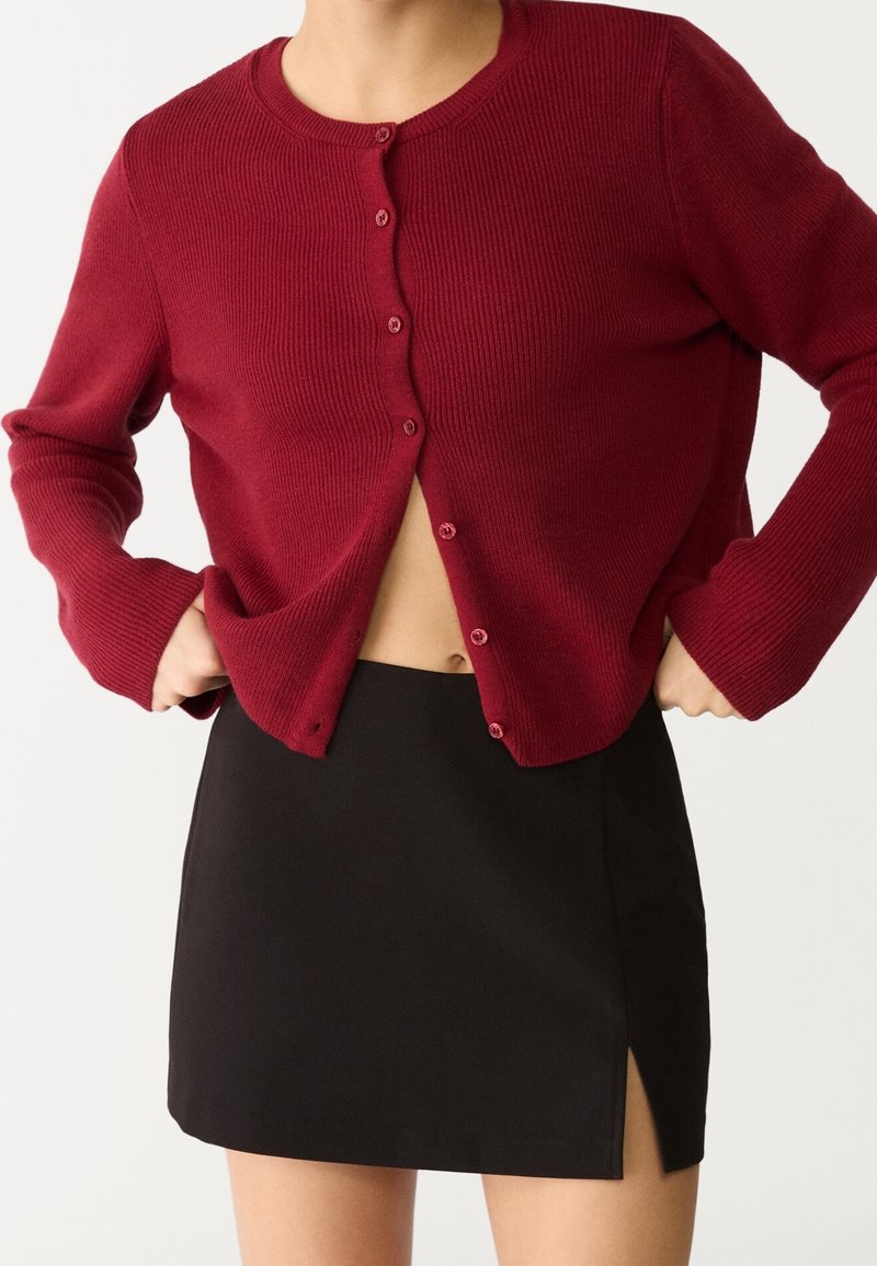 Burgundy ribbed cardigan with round neckline and button closure, paired with a fitted black mini skirt featuring a side slit.