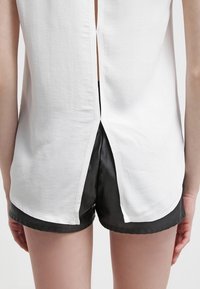 Weißes Oberteil mit strukturierter Oberfläche und hinten Schlitz; kombiniert mit schwarzen Satinshorts, die die kontrastierenden Materialien und Farben betonen.