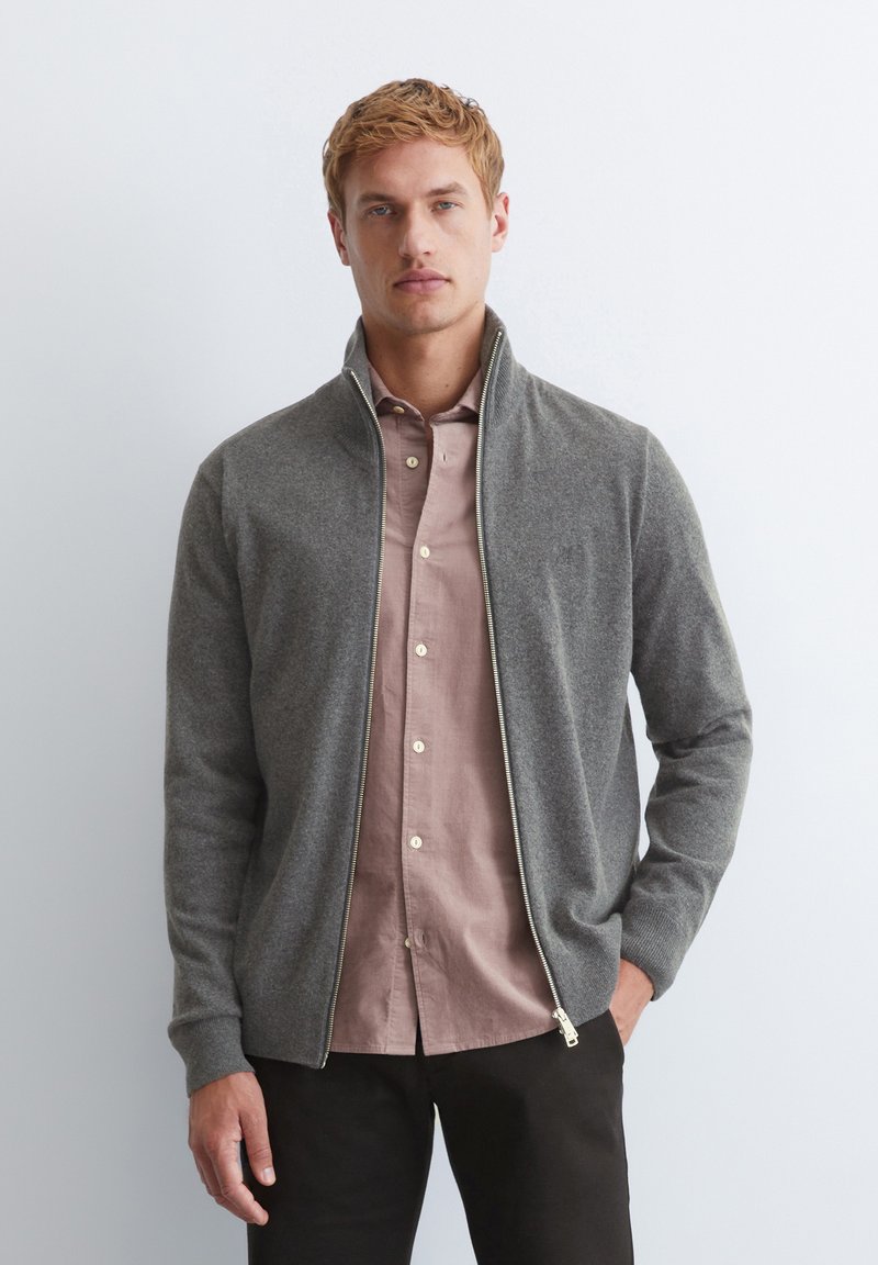 Gray zip-up sweater with a collar, featuring ribbed cuffs. Layered over a light pink button-up shirt, shown against a plain backdrop.