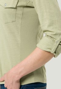 Camicia verde chiaro a maniche lunghe con texture a costine sui lati, polsini arrotolati e una piccola tasca sul petto. Rifinitura del tessuto liscia.