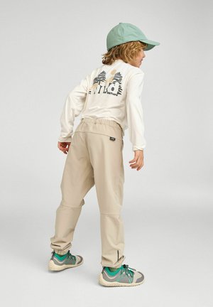 Dziecko ubrane w beżowe spodnie, zielono-szare trampki, białą koszulkę z długim rękawem z grafiką drzewa i słońca oraz zieloną czapkę z daszkiem, stojące bokiem.