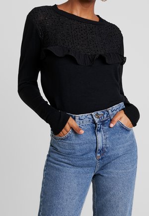 Femme portant un haut noir à manches longues avec panneau avant texturé et détail volanté, associé à un jean bleu taille haute, mains dans les poches.