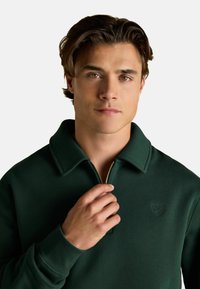 Ciemnozielona rozpinana bluza wykonana z miękkiego materiału, z kołnierzykiem i wytłoczonym logo na klatce piersiowej. Widoczny złoty zamek.