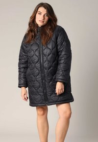 Manteau matelassé noir avec fermeture éclair, encolure arrondie et coupe ample. Présente des coutures en motif circulaire et des manches larges.