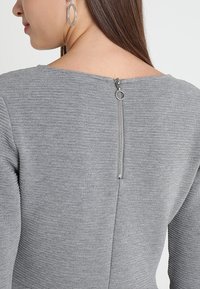 Femme portant un haut gris côtelé avec une fermeture éclair argentée verticale dans le haut du dos, assorti à de grandes boucles d'oreilles argentées en forme de créoles.