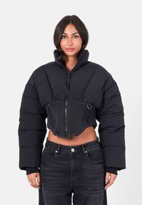 Femme aux cheveux noirs portant une veste doudoune noire courte avec un col haut et un jean noir, se tenant devant un fond blanc uni.