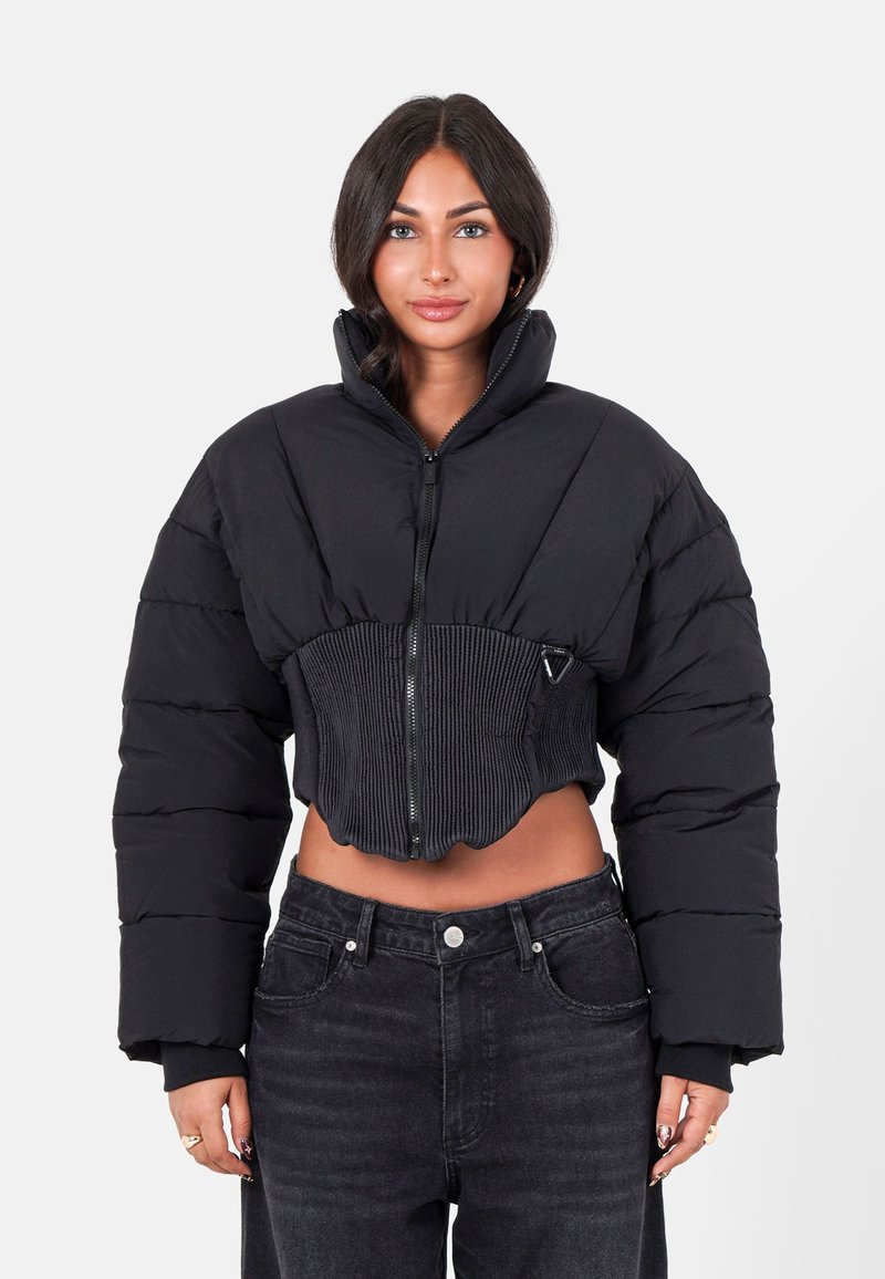 Femme aux cheveux noirs portant une veste doudoune noire courte avec un col haut et un jean noir, se tenant devant un fond blanc uni.