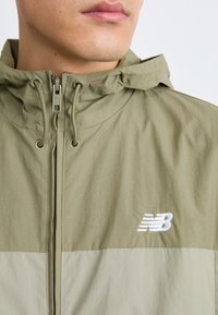 Veste légère couleur vert olive avec capuche. Elle dispose d'une fermeture éclair sur le devant et d'un logo New Balance sur la poitrine. Texture lisse avec un panneau inférieur contrastant.