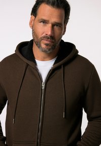 Brun zip-up hættetrøje med en blød, tekstureret sort inderforing, snore og metallynlås. Bæres over en hvid t-shirt.