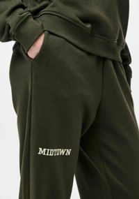 Olive zielone spodnie dresowe o luźnym kroju, miękkiej tkaninie, bocznych kieszeniach oraz białym haftowanym logo "MIDTOWN" na prawej nodze.