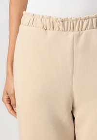 Beige elastic-band broeken met een gladde textuur. Kenmerken zijn een geplooide tailleband en een aangesloten pijpenontwerp, gedeeltelijk verborgen door een hand.