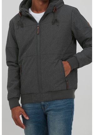 Homme portant une veste à capuche gris foncé zippée avec cordons et un jean bleu, main dans la poche de la veste, sur fond uni.
