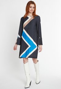 Marineblaues Kleid mit geometrischen Streifen in Blau, Beige und Weiß. Lange Ärmel, lockere Passform und Detailknöpfe an den Ärmelbündchen. Kombiniert mit weißen Stiefeln.