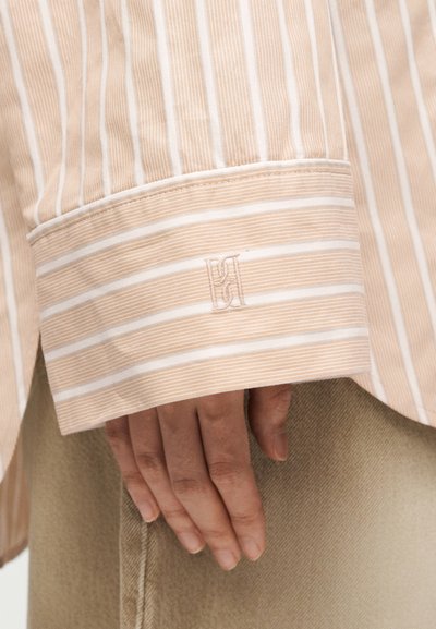 Main reposant près du poignet d'une chemise rayée beige et blanche avec logo brodé, sur fond de pantalon beige clair.