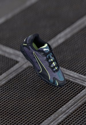 Dunkelgrauer und schwarzer Puma-Sneaker mit neongelben Akzenten, der auf einem strukturierten Metallrost liegt.