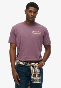 Camiseta de algodón morada de manga corta, con el logo "Superdry Los Ángeles" en color crema. Pantalones vaqueros con una camisa de cuadros atada a la cintura.