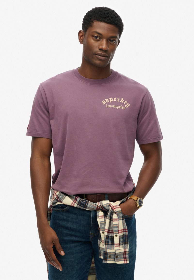 Camiseta de algodón morada de manga corta, con el logo "Superdry Los Ángeles" en color crema. Pantalones vaqueros con una camisa de cuadros atada a la cintura.