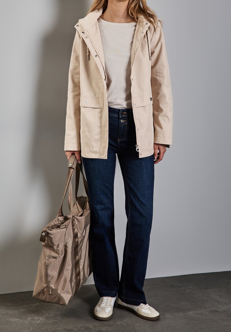 Ljusbeige huva-jacka med silverdragkedja, två framfickor, ihop med mörkblå jeans och vita sneakers. Bär en brun tote-väska.