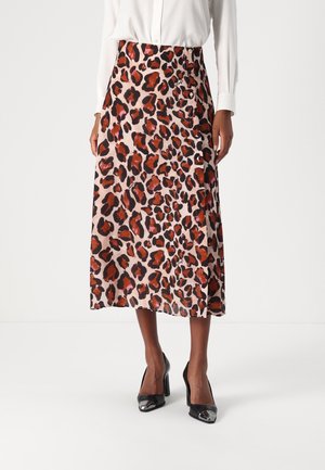 Femme portant une jupe midi imprimé animal rose et marron, un chemisier blanc à manches longues et des chaussures à talons noirs, debout sur un sol blanc.