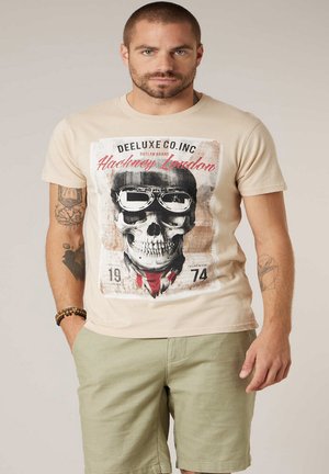 Man met tatoeages, gekleed in een beige grafisch T-shirt met een ontwerp van een schedel met een bril en lichtgroene korte broek, staand met één hand in de zak.