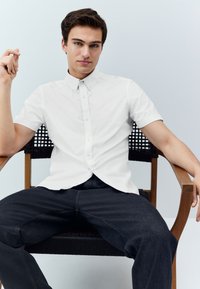 Chemise blanche à manches courtes avec col et boutons, associée à un jean en denim foncé. Le modèle est assis sur une chaise en bois avec un dossier tressé.