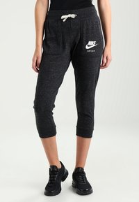 Šedé zkrácené tepláky s dvěma bočními kapsami, stahovací šňůrkou v pase a bílým logem Nike na levém stehně. Nosené s černými teniskami.