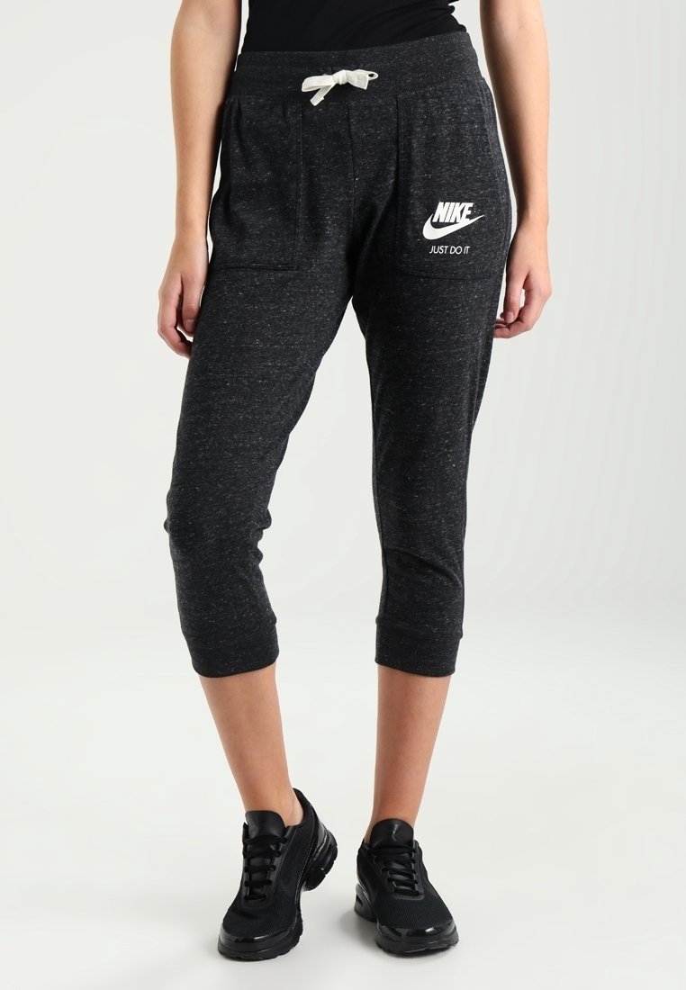 Šedé zkrácené tepláky s dvěma bočními kapsami, stahovací šňůrkou v pase a bílým logem Nike na levém stehně. Nosené s černými teniskami.