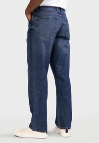 Μπλε τζιν με ίσια εφαρμογή, με δύο πίσω τσέπες, πορτοκαλί ραφές και ελαφρώς ξεθωριασμένη υφή. Φοριέται με λευκά αθλητικά παπούτσια.