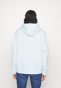 Sudadera con capucha de color azul claro, con una capucha grande, puños y dobladillo acanalados, y una textura suave. Llevada con vaqueros azules, vista desde atrás.