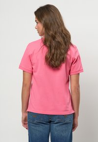 Ženska z dolgimi rjavimi lasmi, oblečena v roza majico s kratkimi rokavi in modre kavbojke, stoji obrnjena stran proti enostavni ozadju.