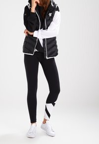 Černobílá péřová vesta s kapucí, doplněná černými legínami s bílým geometrickým vzorem a bílými teniskami.