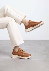 Zapatillas de cuero marrón con suela de goma beige, con cordones y costuras de acento, combinadas con pantalones acampanados de color claro.