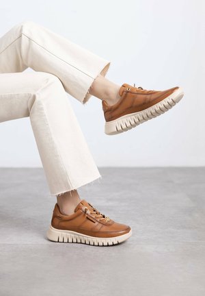 Zapatillas de cuero marrón con suela de goma beige, con cordones y costuras de acento, combinadas con pantalones acampanados de color claro.