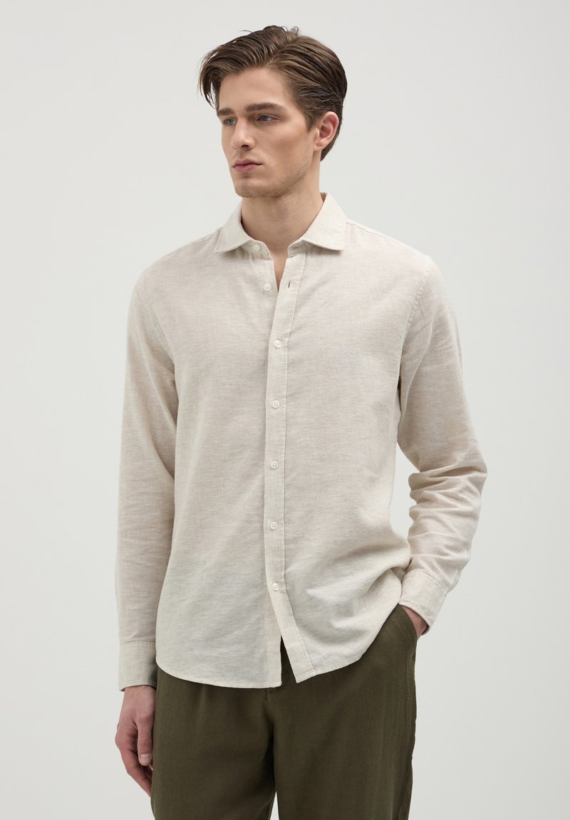 Beige Leinen-Hemd mit langen Ärmeln, klassischem Kragen und Knopfverschluss. Es hat einen leicht strukturierten Stoff und eine slim fit Passform und wird mit olivgrünen Hosen kombiniert.
