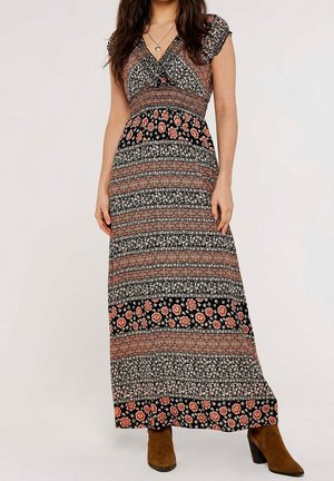 Femme portant une robe longue noire et rouille à motifs floraux et géométriques, avec des manches courtes et des bottines en daim marron.