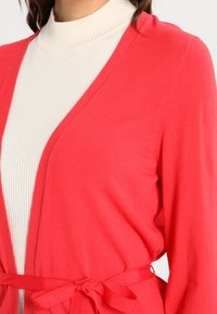 Cárdigan de punto coral con cinturón, con escote en V y mangas largas, superpuesto a un cuello de tortuga blanco de canalé. Textura suave, ligero.