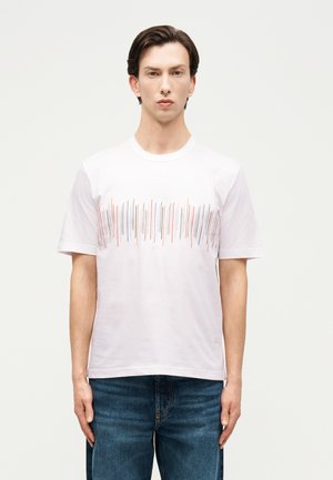 Jonge man met een wit T-shirt met meerkleurige verticale strepen over de borst en een blauwe spijkerbroek, staand tegen een effen achtergrond.