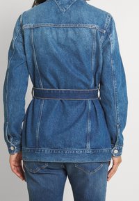 Chaqueta de mezclilla en azul oscuro, con una cintura ceñida por cinturón, mangas largas y un cuello clásico. Costuras visibles y bolsillos laterales.