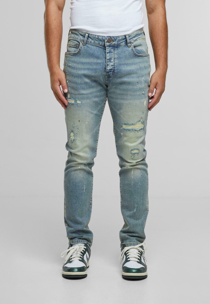 Persona con jeans desgastados de color azul claro con manchas de pintura y zapatillas oscuras verdes y blancas, de pie contra un fondo liso.