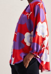 Rotes und lila langärmeliges Oberteil mit großem Blumenmuster, elastischen Bündchen und Seitenschlitzen. Glatter Stoff mit lässiger Silhouette.