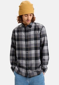 Gray plaid shirt with black and light gray squares, yellow accents, button-up design, and a chest pocket. Model wears a mustard knit beanie.